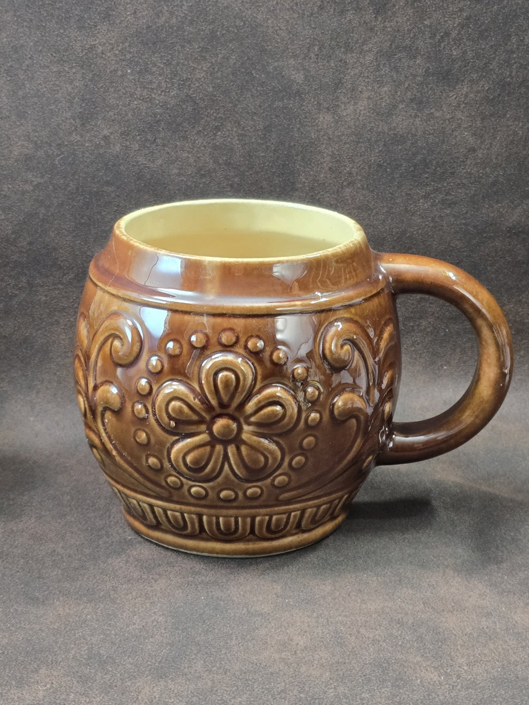 Vintage Keramikbecher mit brauner Glasur, floralem Reliefmuster und hellem Innenbereich in klarer Frontalansicht auf dunklem Hintergrund