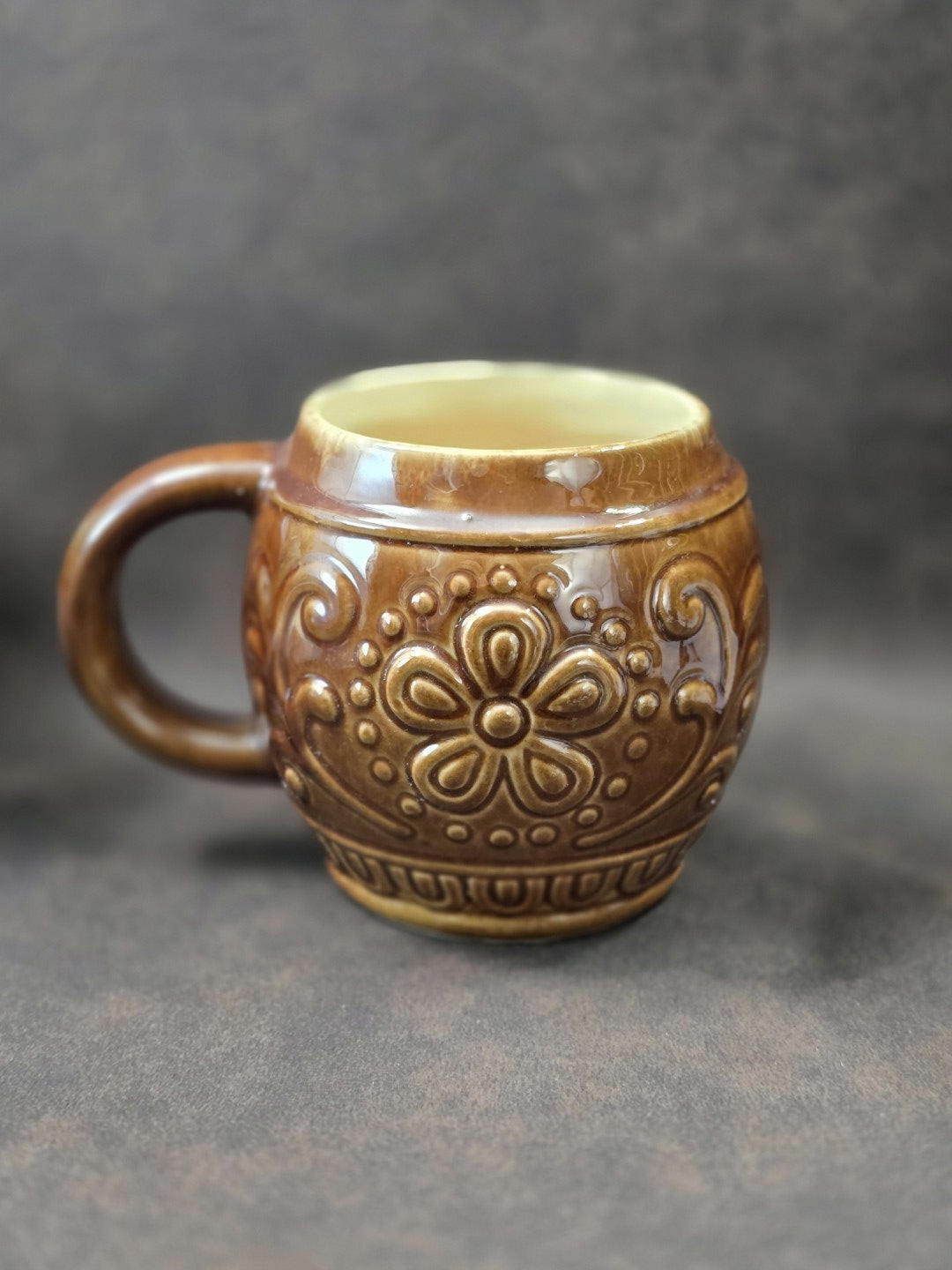 Vintage Keramikbecher mit brauner Glasur, symmetrischem Blumenrelief und seitlichem Henkel in gedrehter Ansicht auf dunklem Hintergrund