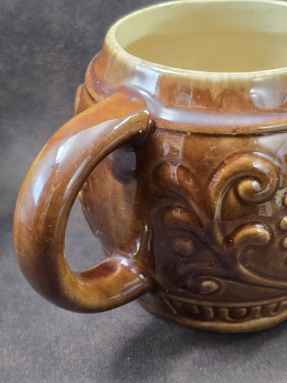 Vintage Keramikbecher mit braun glasiertem Henkel, hellem Innenbereich und reliefiertem Korpus in enger Nahaufnahme der Henkelpartie