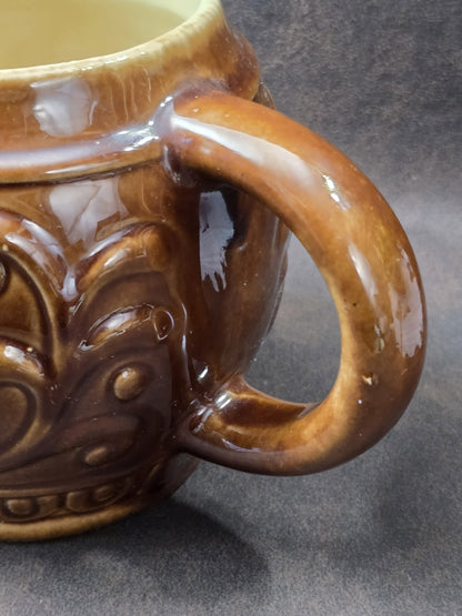 Vintage Keramikbecher mit braun glasiertem Henkel, hellem Innenbereich und reliefiertem Korpus in enger Nahaufnahme der Henkelpartie