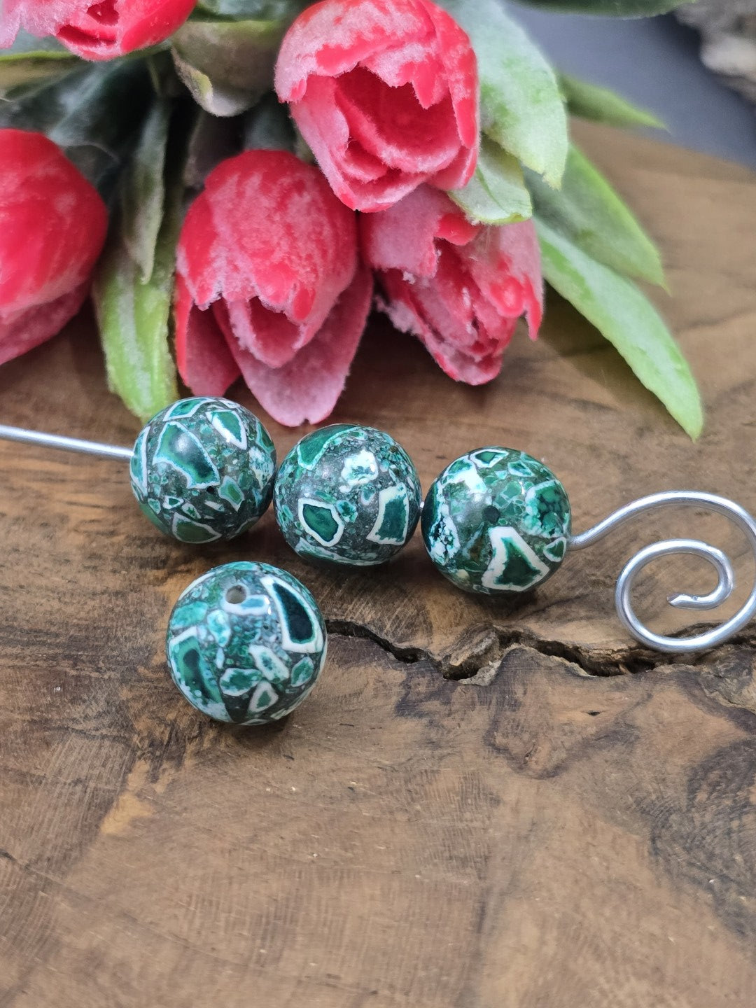 Grüne Mosaik-Steinperlen (12 mm Kugel) auf Draht aufgereiht vor dekorativen roten Blütendetails.