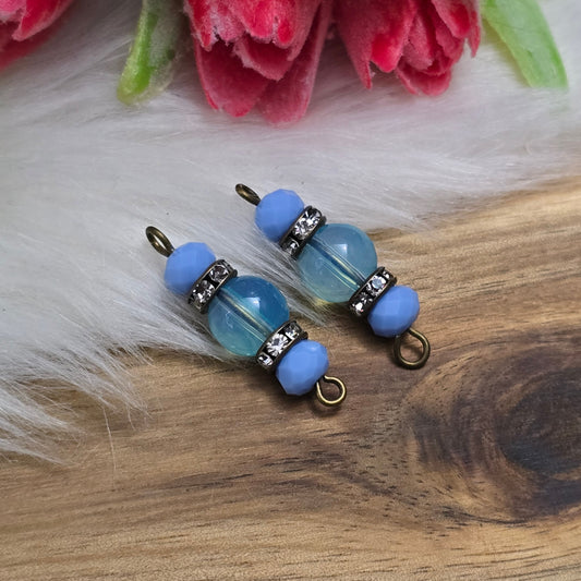 Das Bild zeigt zwei handgefertigte Verbinder mit hellblauen Glasperlen. Die Kombination aus einer transparenten hellblauen Glasperle (8mm) und einer opaken hellblauen Kristallglasperle wird durch ein funkelndes Strassrondell ergänzt. Antik bronzefarbene Metallelemente verleihen den Verbindern einen eleganten Vintage-Look. Mit einer Länge von 27mm sind sie ideal für die Schmuckherstellung.