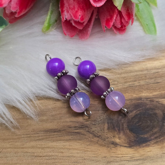Das Bild zeigt zwei handgefertigte Verbinder mit lila und violetten Glasperlen in unterschiedlichen Ausführungen – eine opake, eine matte und eine milchig transparente. Die Perlen sind harmonisch angeordnet und werden von detailreich verzierten, antik-silberfarbenen Metallelementen eingefasst. Die filigranen Zwischenelemente verleihen den Verbindern eine edle Vintage-Optik. Mit einer Länge von ca. 33mm eignen sie sich perfekt für kreative Schmuckdesigns wie Armbänder, Ketten oder Ohrringe.