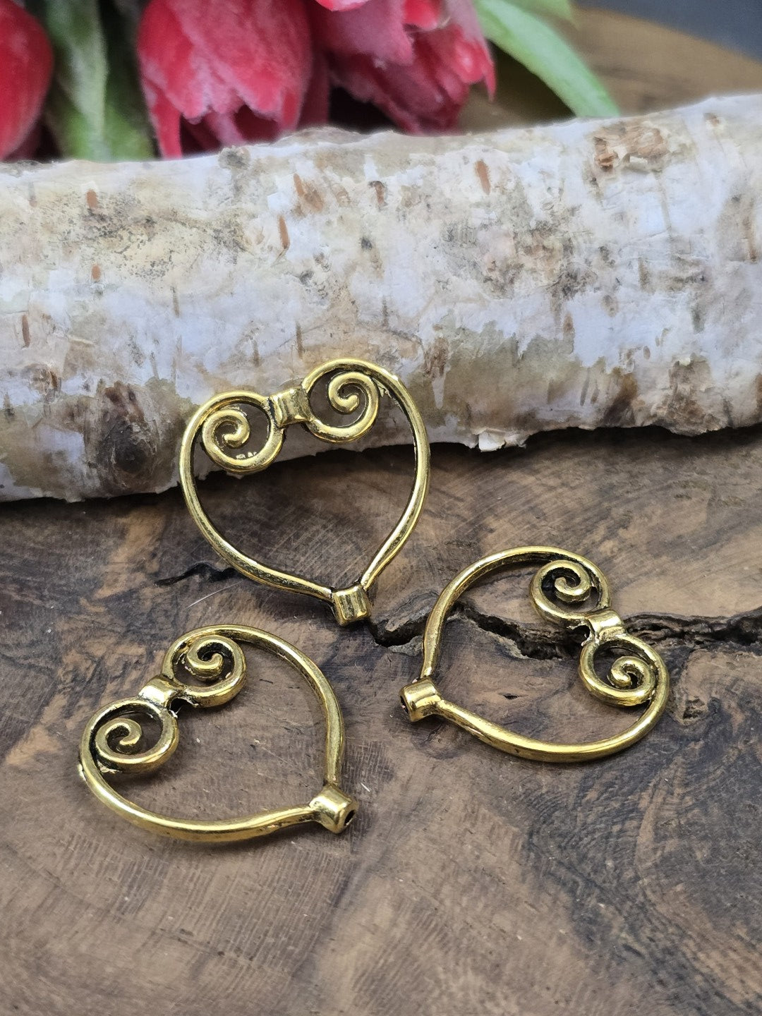 Drei goldfarbene Perlenrahmen in Herzform (21 mm) mit filigranem Schnörkel-Muster flach liegend auf Holzgrund vor Birkenrinde.