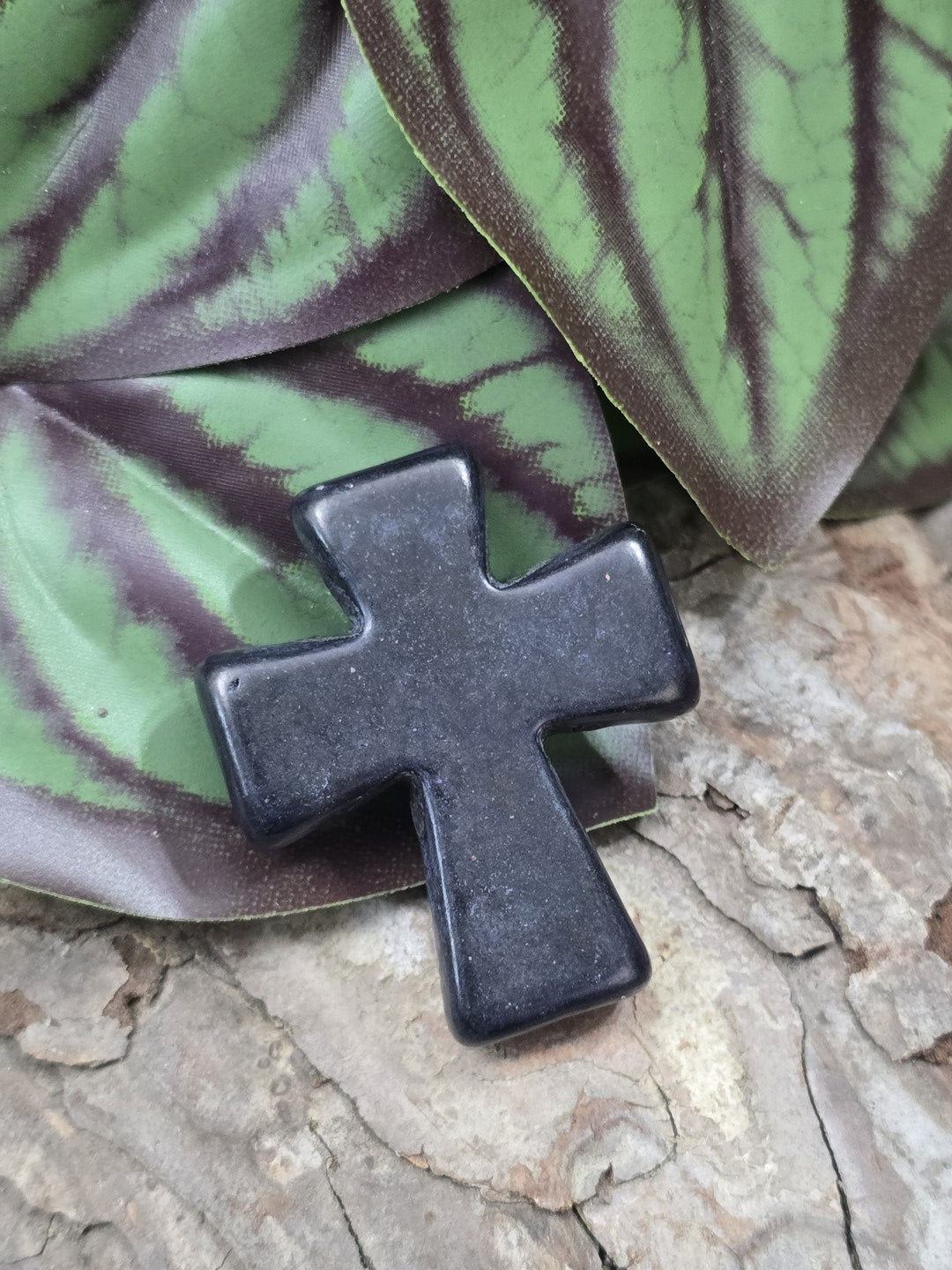 Schwarze Howlith Kreuzperle (36 mm) flach liegend auf rustikaler Baumrinde vor grün gestreiftem Blatt