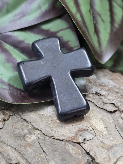 Schwarze Howlith Kreuz-Schmuckperle auf Baumrinde mit gut sichtbarer Bohrung am unteren Ende