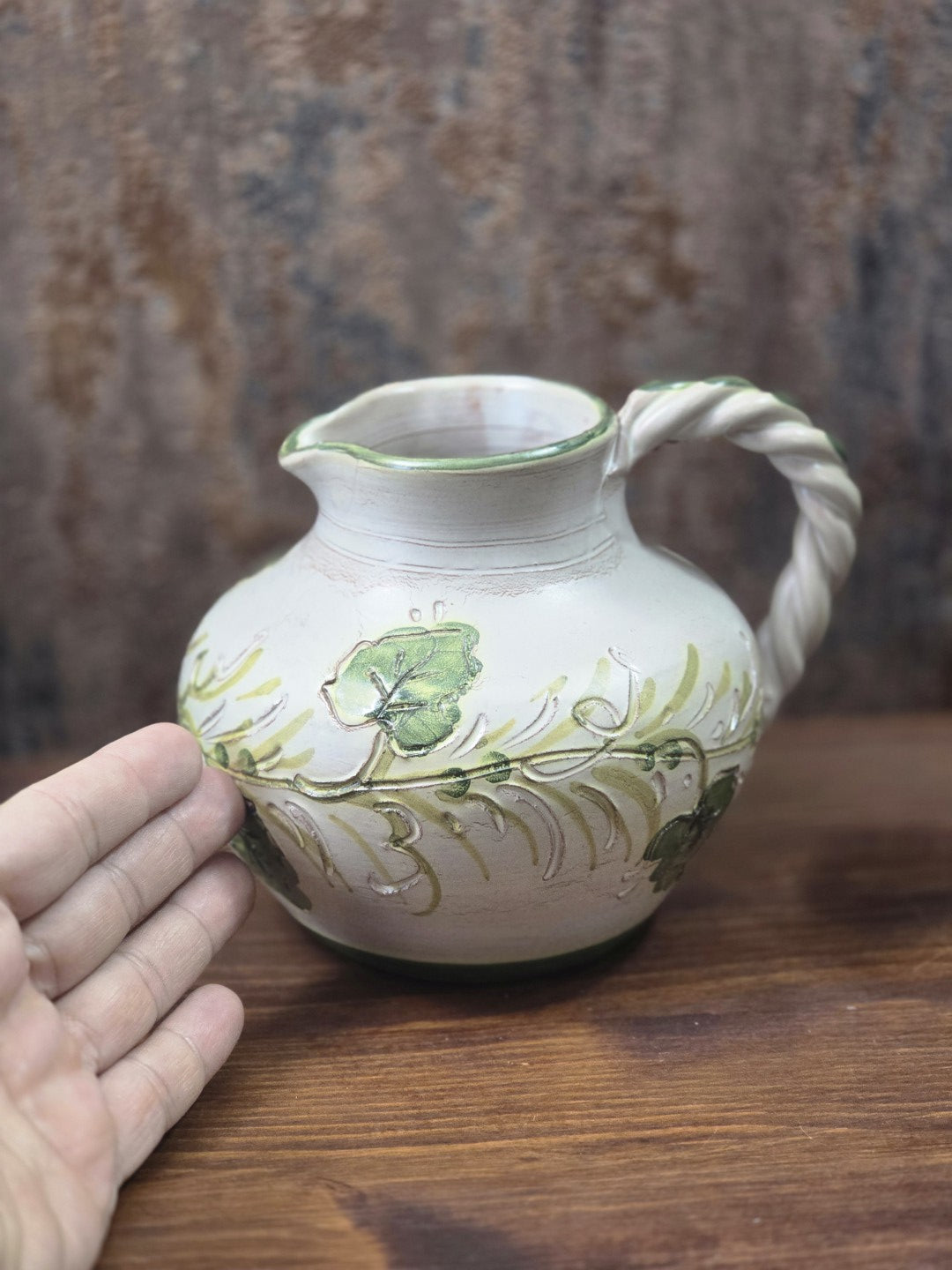 Vintage Keramikkrug mit cremefarbener Oberfläche, grünem Blattdekor und gedrehtem Henkel neben einer Hand als Größenvergleich