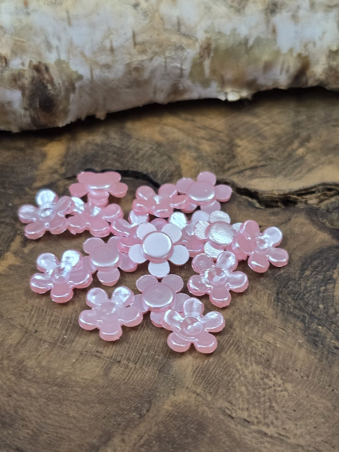 Eine Gruppe kleiner, rosafarbener Klebeblumen (8 mm) mit perlmuttartig schimmernder Oberfläche und flacher Rückseite auf einer rustikalen Holzscheibe.