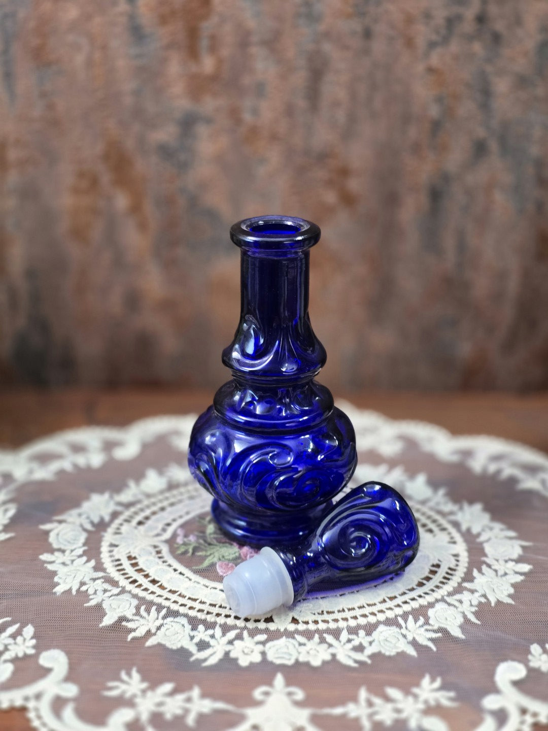Vintage Glasflakon in Kobaltblau mit geöffnetem Flaschenhals und separat liegendem Glasstopfen mit weißem Dichteinsatz auf Spitzendecke