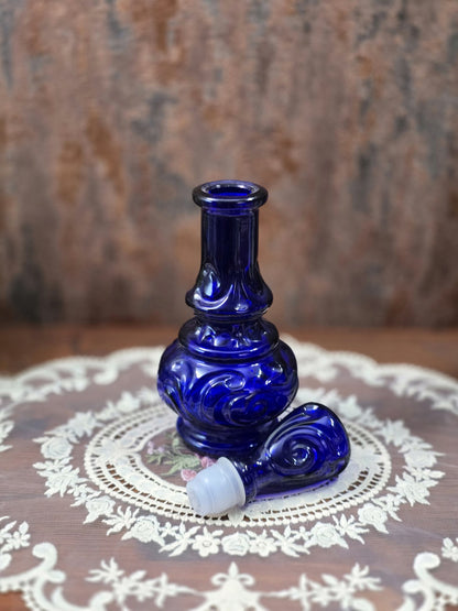 Vintage Glasflakon in Kobaltblau mit geöffnetem Flaschenhals und separat liegendem Glasstopfen mit weißem Dichteinsatz auf Spitzendecke