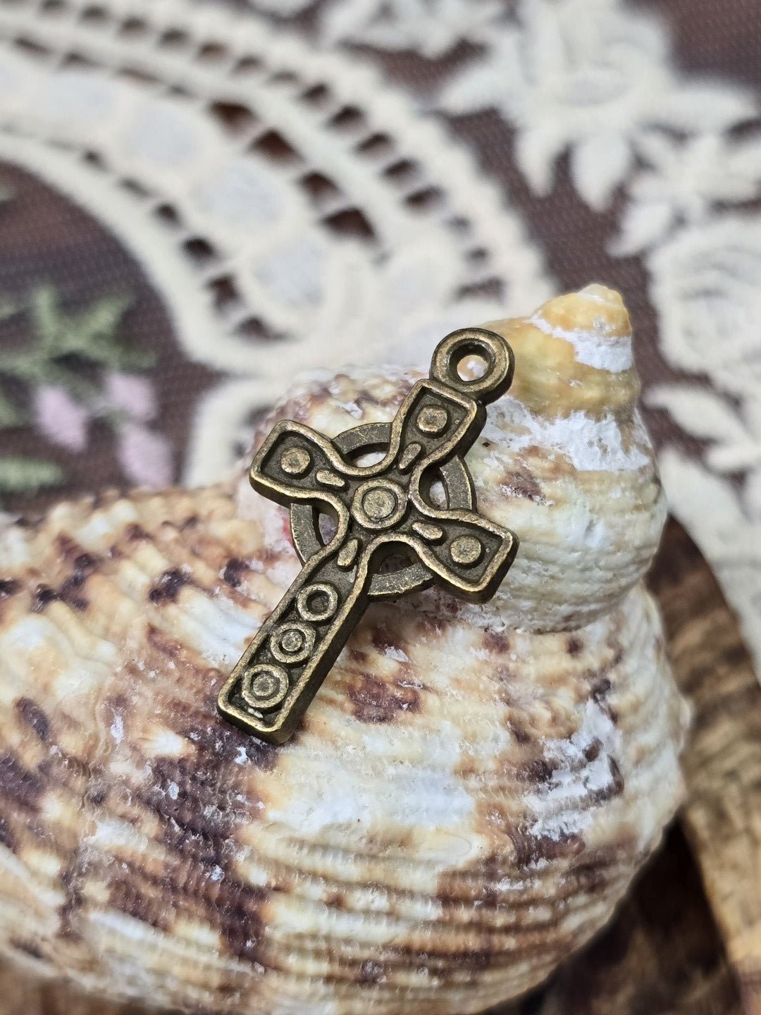 Keltenkreuz-Schmuckanhänger in Antikbronze mit Kreis-Muster aufgestellt vor einer Muschel.
