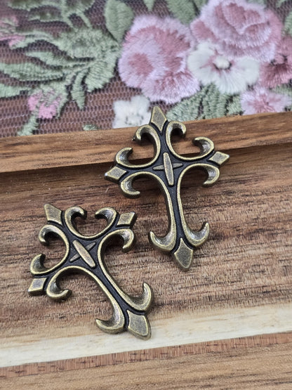 Zwei barocke Kreuz-Anhänger in Antikbronze flach liegend auf Holzgrund.