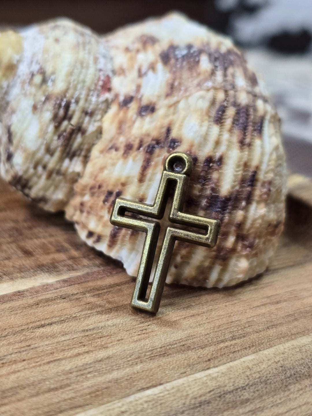 Kreuz-Charm in Antikbronze mit Vertiefung angelehnt an eine Muschel auf Holz für Schmuckdesign.