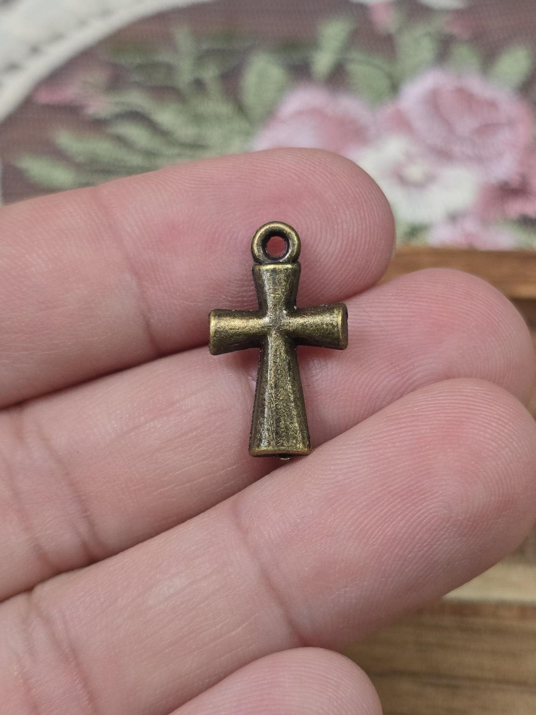 Kleine Kreuz-Perle 20x12mm in Bronze-Optik auf zwei Fingern gehalten als Größenvergleich.