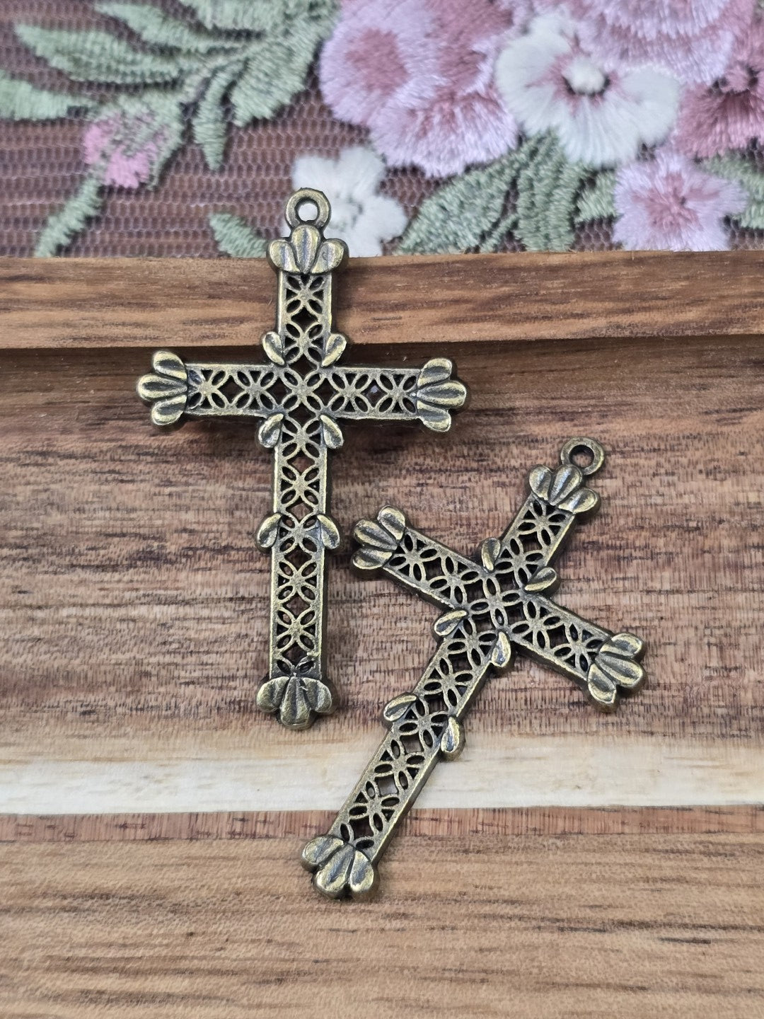Zwei filigrane Kreuz-Anhänger in Antikbronze mit Lochmuster flach liegend auf Holzgrund.