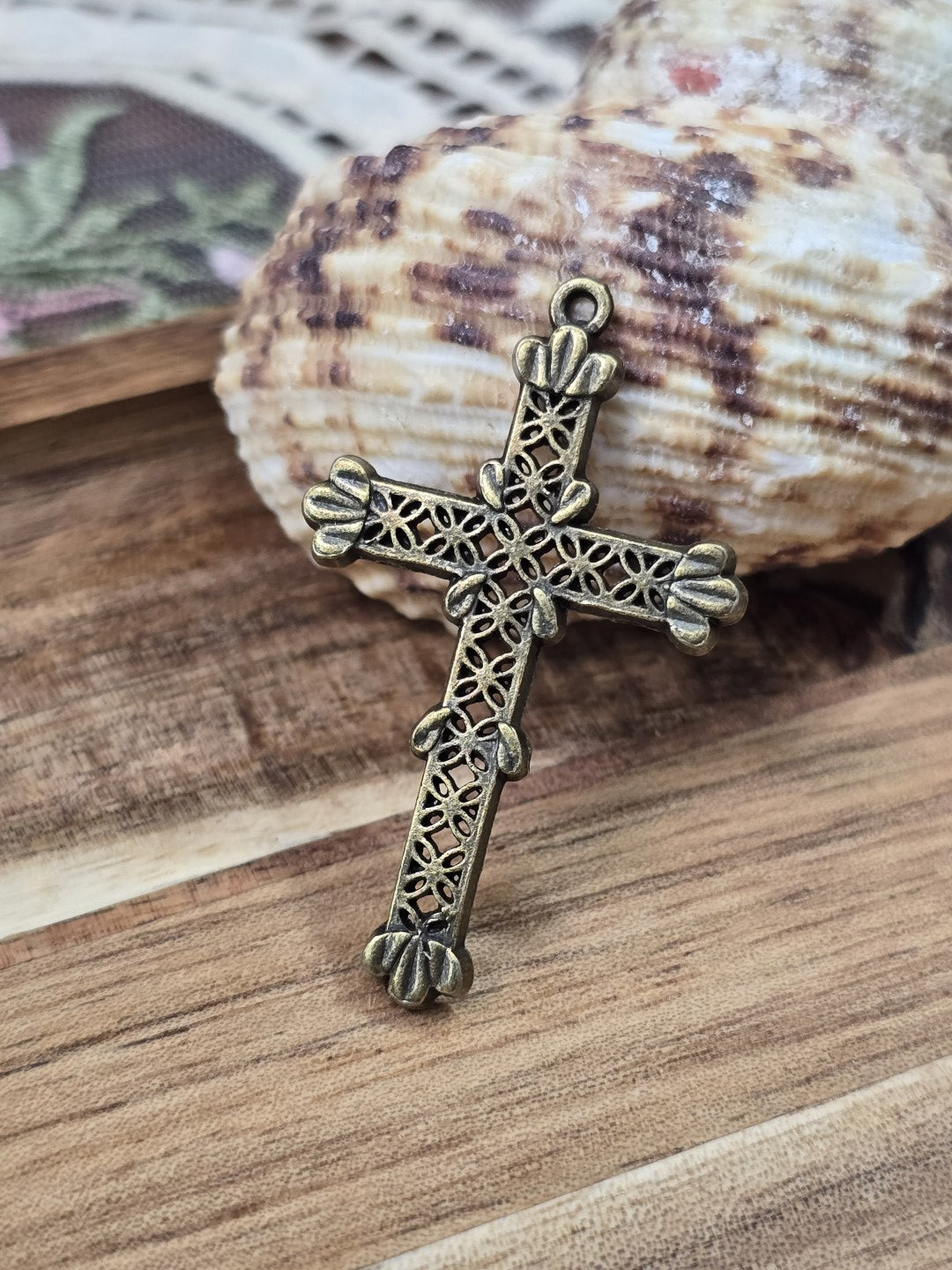Verzierter Vintage-Kreuz-Schmuckanhänger mit Öse aufgestellt vor einer dekorativen Muschel.