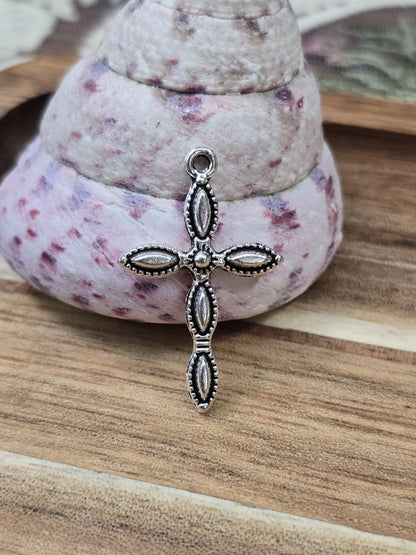 Verzierter Kreuz-Charm in Silber-Optik stehend vor dekorativer Muschel für Schmuckherstellung.