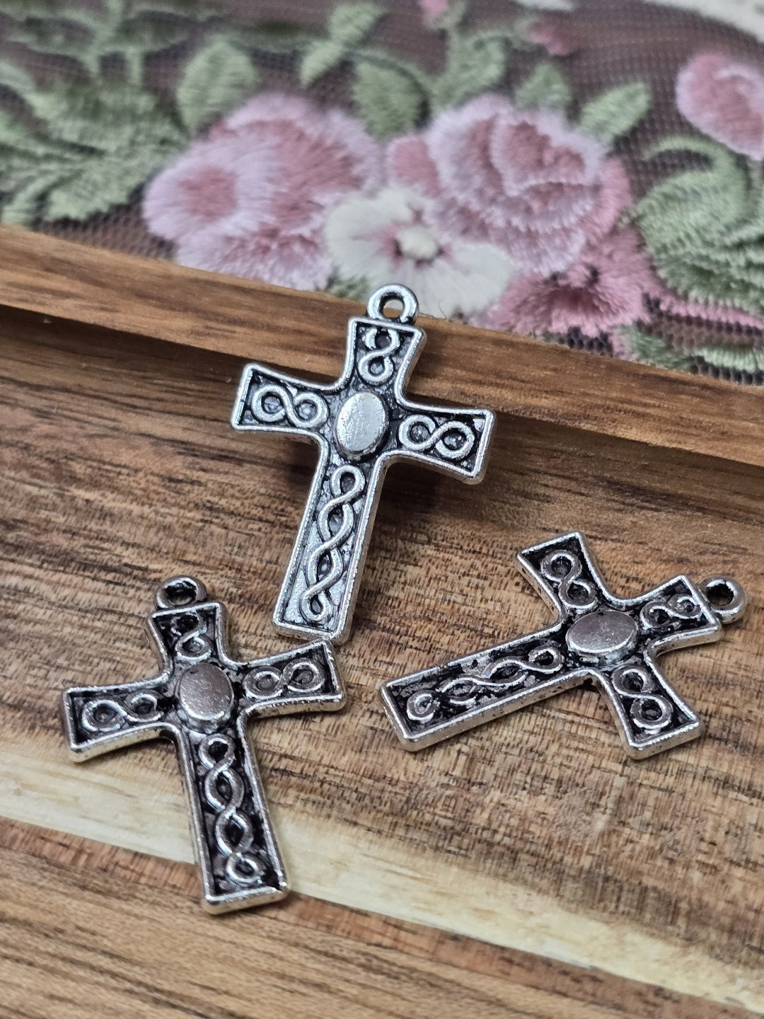 Drei kleine Kreuz-Anhänger in Antiksilber mit keltischem Flechtmuster und Öse flach liegend auf Holzgrund.