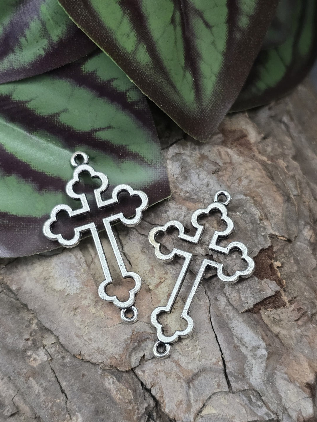 Zwei filigrane Kreuz-Verbinder in Antik-Silber (35 mm) mit Kleeblatt-Enden flach liegend auf rustikalem Holzgrund vor grünem Blattwerk.