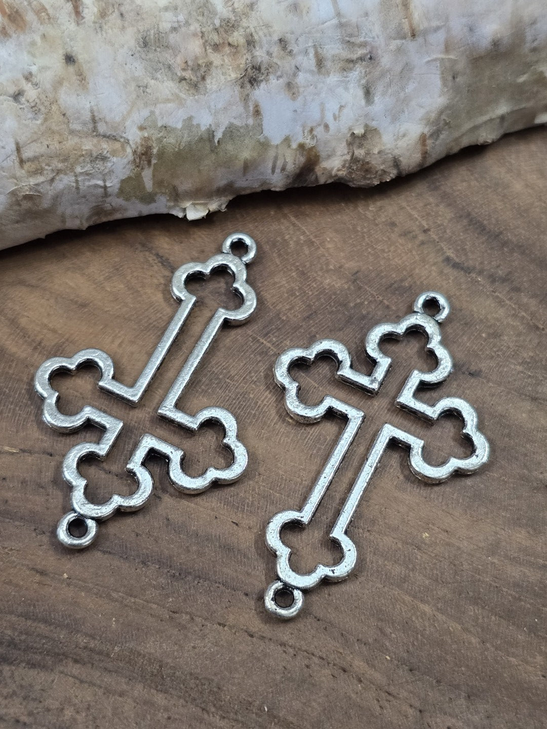 Zwei filigrane Kleeblatt-Kreuz-Verbinder (35 mm) in Antik-Silber flach nebeneinander liegend auf einer Holzplatte.