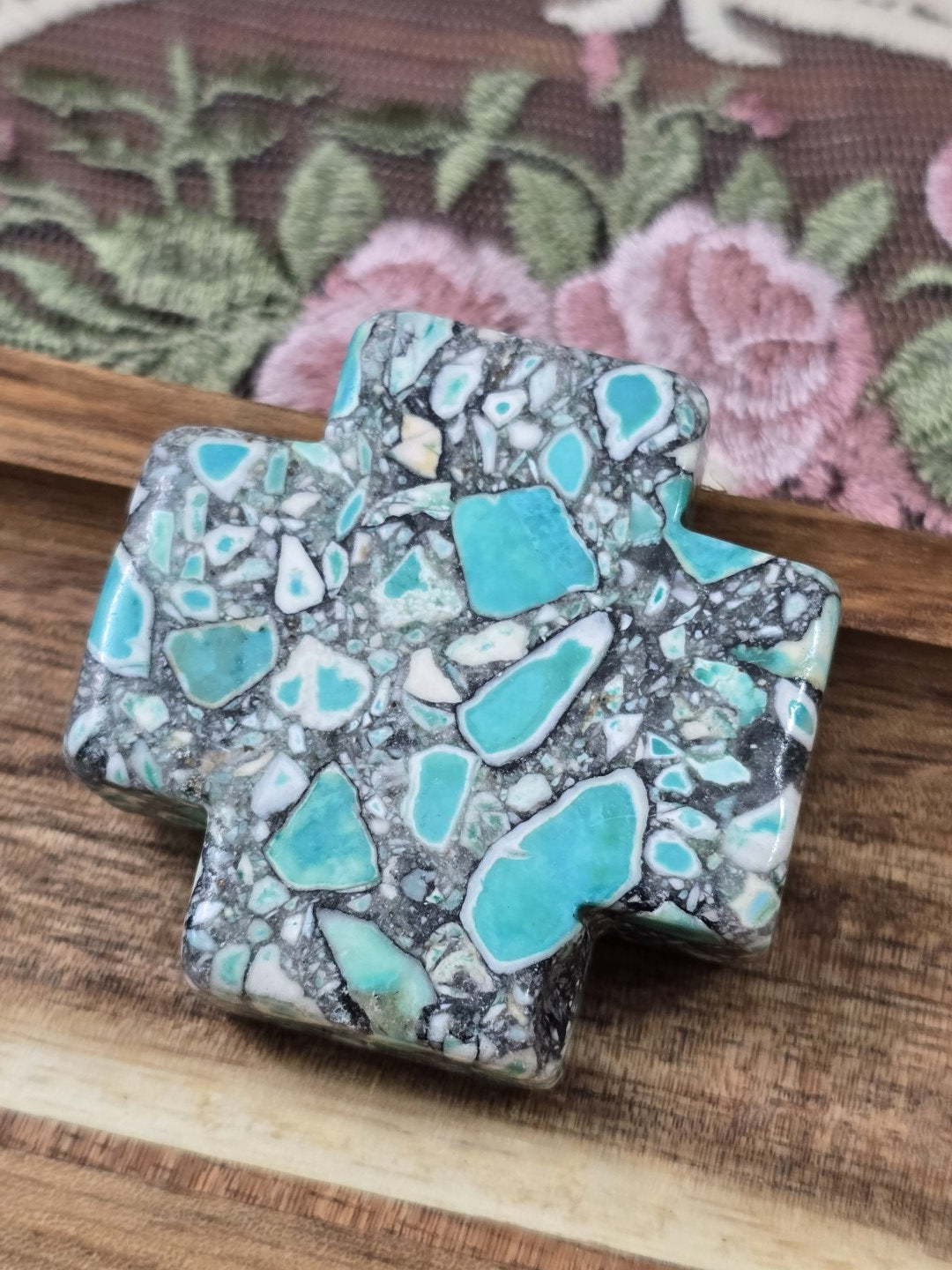 Mosaik-Kreuzperle aus Stein-Komposit mit türkis-grünen Akzenten auf Holzplatte.