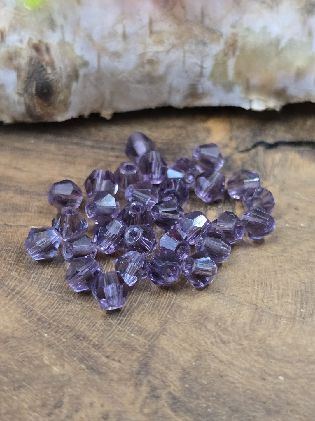 Ansammlung von 30 lilafarbenen Kristallglas-Bicones (4 mm Doppelkegelperlen) auf rustikalem Holzgrund.