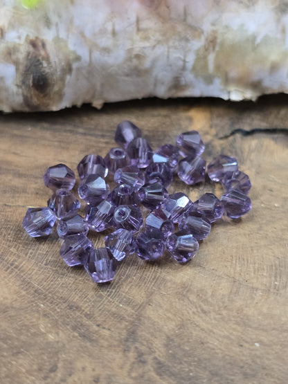 Ansammlung von 30 lilafarbenen Kristallglas-Bicones (4 mm Doppelkegelperlen) auf rustikalem Holzgrund.