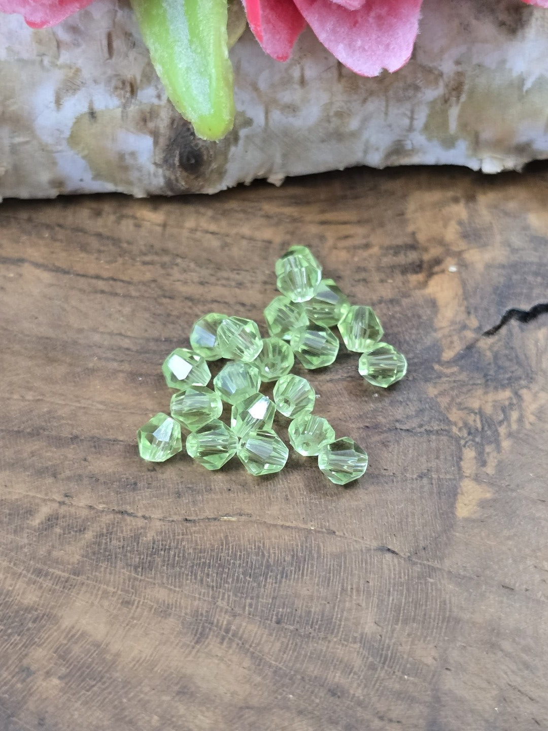 Ansammlung von 30 peridotgrünen Kristallglas-Bicones (4 mm Doppelkegelperlen) auf rustikalem Holzgrund.