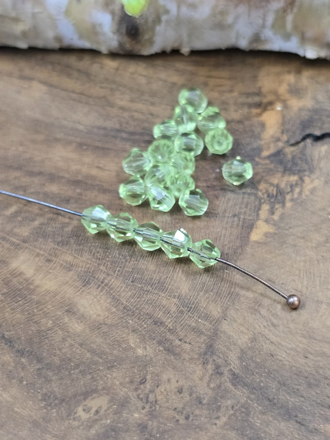 Sechs peridotgrüne Kristallglas-Bicones (4 mm) auf einen Nietstift aufgereiht neben einem Haufen loser Glasperlen.