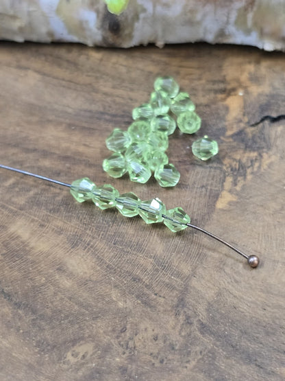Sechs peridotgrüne Kristallglas-Bicones (4 mm) auf einen Nietstift aufgereiht neben einem Haufen loser Glasperlen.