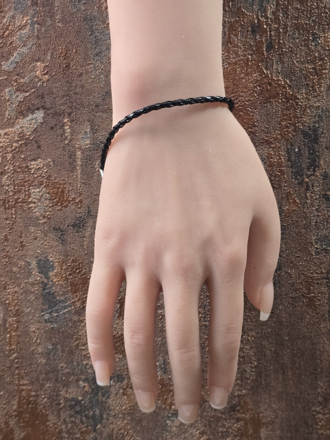 Geflochtenes schwarzes Kunstlederarmband getragen an einer Schaufensterpuppen-Hand vor rustikalem Hintergrund.
