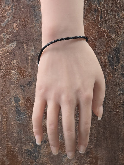 Geflochtenes schwarzes Kunstlederarmband getragen an einer Schaufensterpuppen-Hand vor rustikalem Hintergrund.