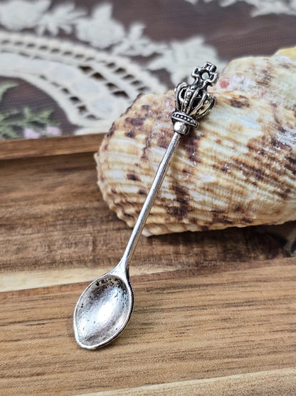 Löffel-Anhänger in Silber-Optik mit Krone dekoriert vor einer Muschel auf Holzgrund.