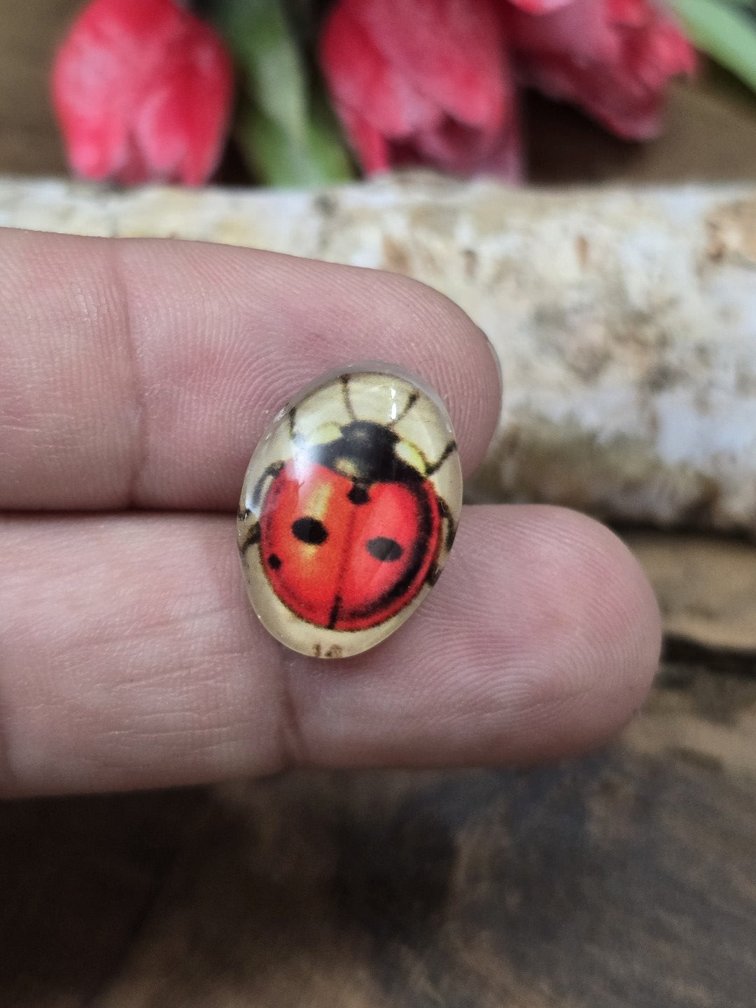 Ein ovaler Marienkäfer-Glascabochon (18x13 mm) in Rot und Schwarz auf einer Fingerspitze als praktischer Größenvergleich für DIY-Schmuck.