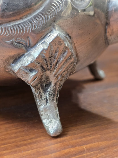 Versilberte Teekanne mit Detail am Standfuß, Muscheldekor, dunkler Patina und Holzuntergrund im Hintergrund