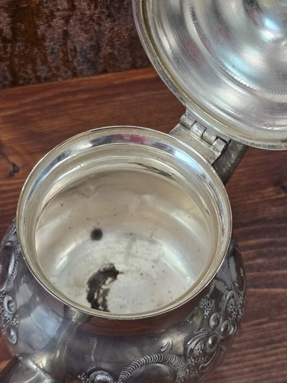 Versilberte Teekanne als Detailaufnahme mit geöffneter Innenfläche, dunklen Rückständen und floral geprägtem Rand