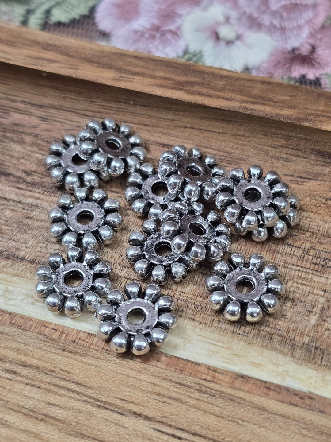 Zwölf silberne Rädchen-Metallperlen in Blütenform auf Holzuntergrund für DIY-Schmuck.