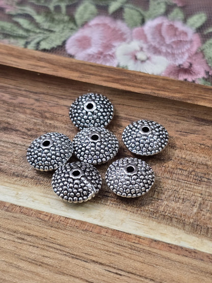 Sechs ufo-förmige Metallperlen in Altsilber mit gepunkteter Kugel-Textur auf Holzgrund.