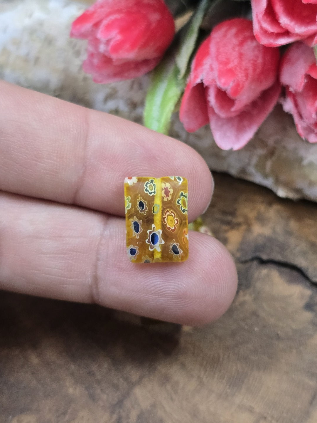 Eine rechteckige gelbe Millefiori-Glasperle mit Blütenmuster auf der Fingerkuppe als Größenvergleich.