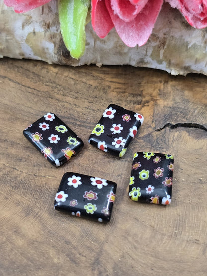 Vier rechteckige schwarze Millefiori-Glasperlen (14x10 mm) mit bunted Blumenmuster flach auf Holzgrund platziert.