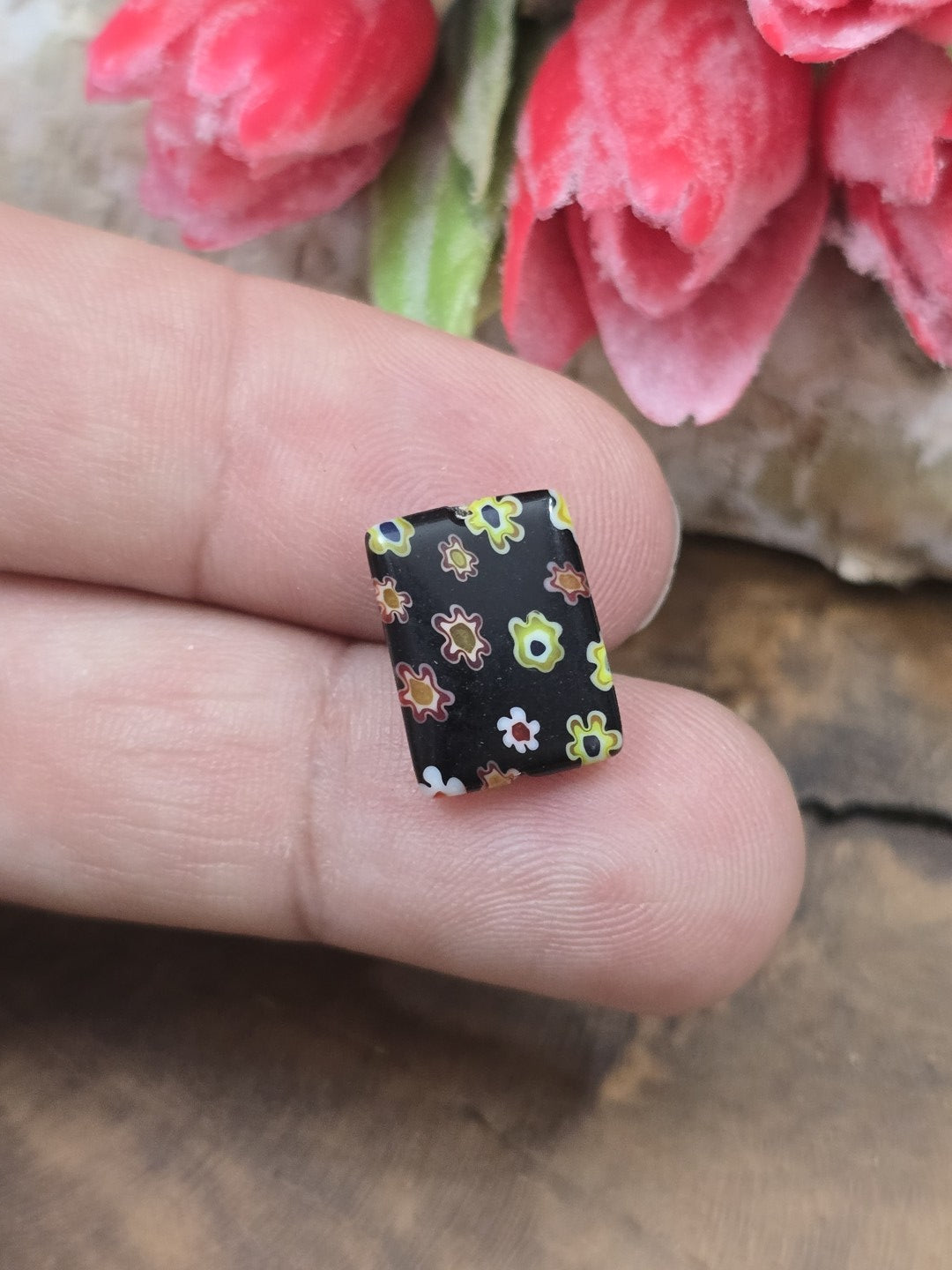 Eine rechteckige schwarze Millefiori-Glasperle mit Blütenmuster auf der Fingerkuppe als Größenvergleich.