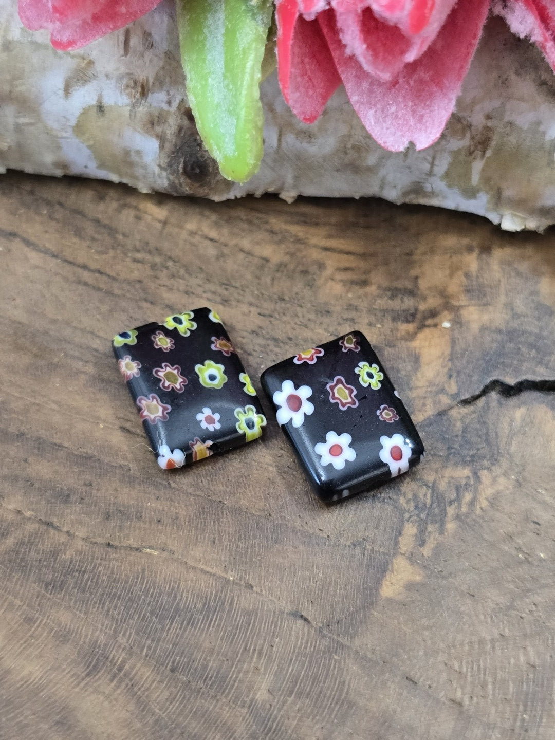 Zwei rechteckige Millefiori-Glasperlen in Schwarz mit bunten Blumenakzenten nebeneinander auf Holz.