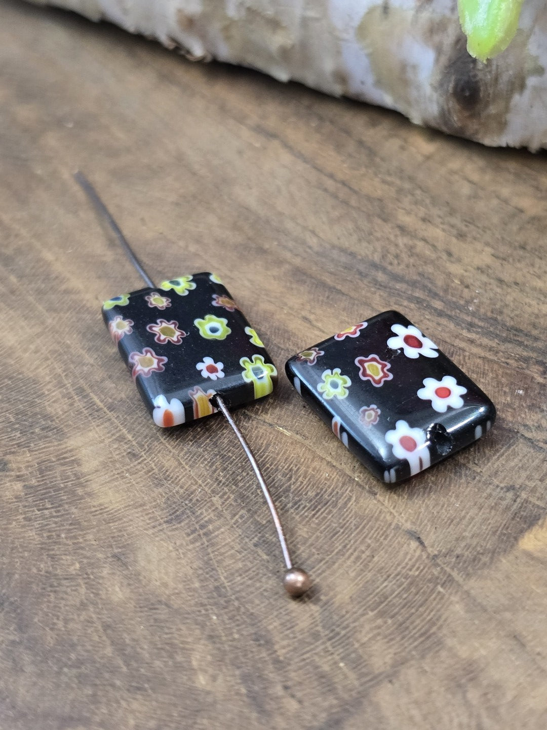 Eine schwarze Millefiori-Glasperle auf einen Nietstift aufgereiht neben einer weiteren Perle zur Veranschaulichung der Bohrung.
