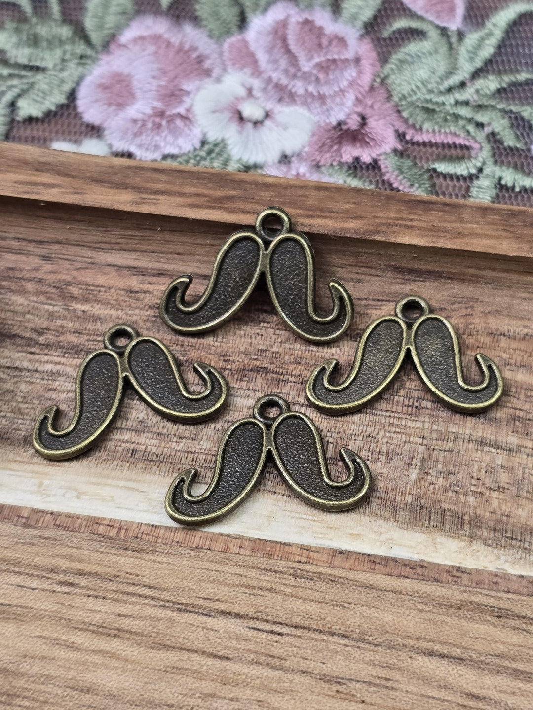 Vier kleine Schnurrbart-Anhänger in Antikbronze mit Öse flach liegend auf Holzgrund.