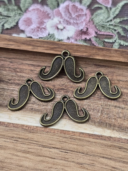 Vier kleine Schnurrbart-Anhänger in Antikbronze mit Öse flach liegend auf Holzgrund.