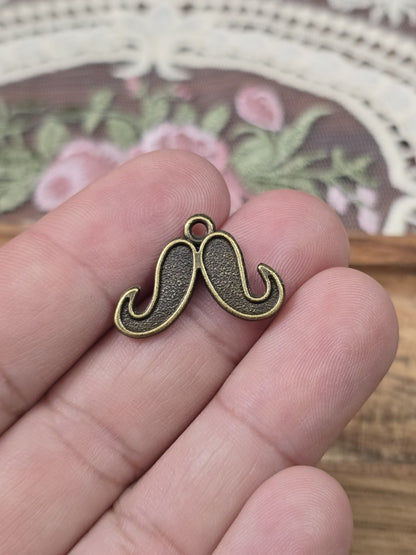 Kleiner Oberlippenbart-Charm 16mm in Bronze-Optik auf einer Hand gehalten als Größenvergleich.