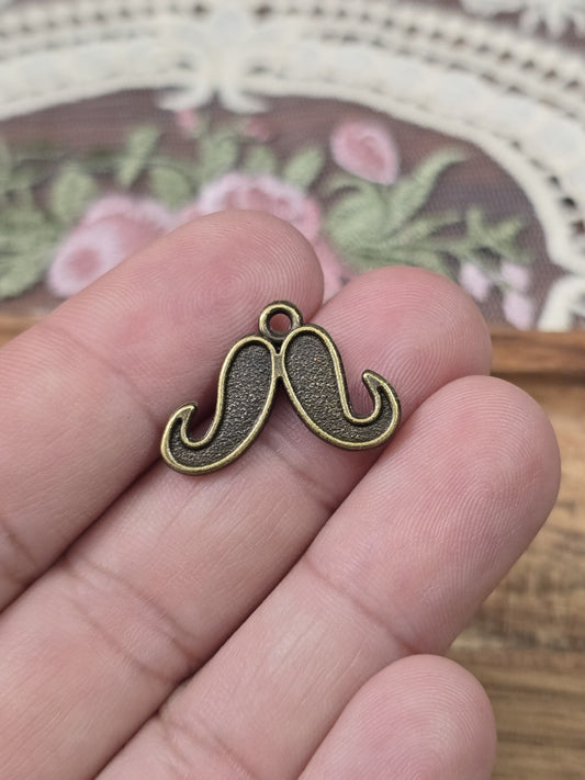 Kleiner Oberlippenbart-Charm 16mm in Bronze-Optik auf einer Hand gehalten als Größenvergleich.