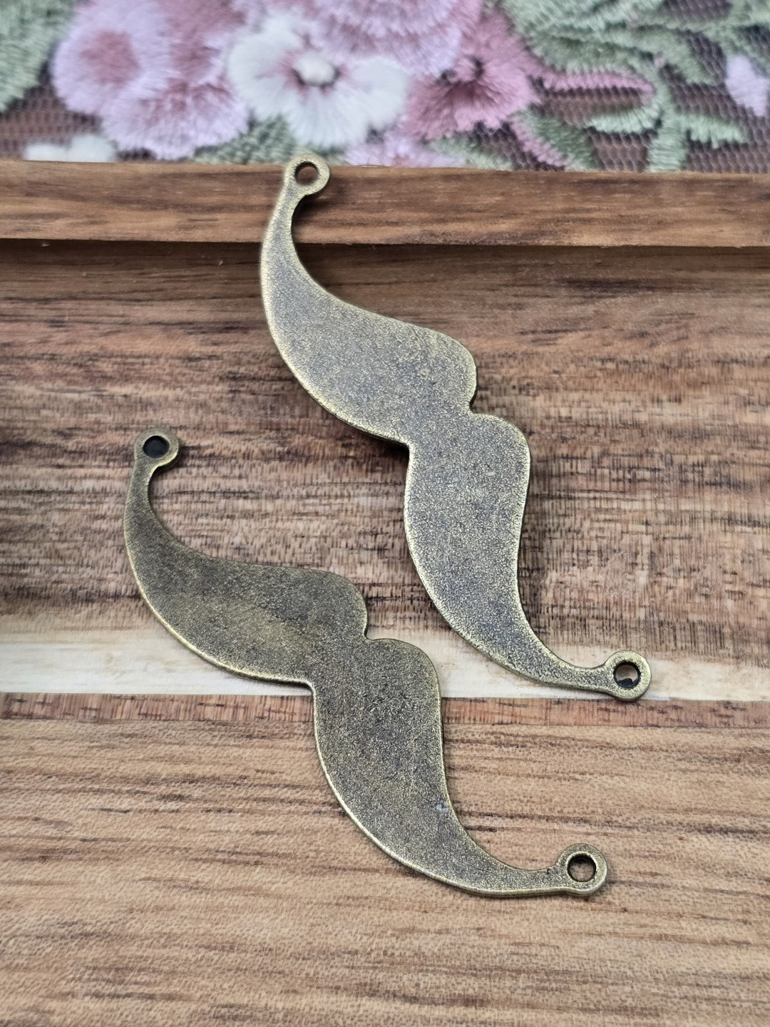 Zwei Schnurrbart-Schmuckverbinder in Antikbronze mit zwei seitlichen Ösen flach liegend auf Holzgrund.