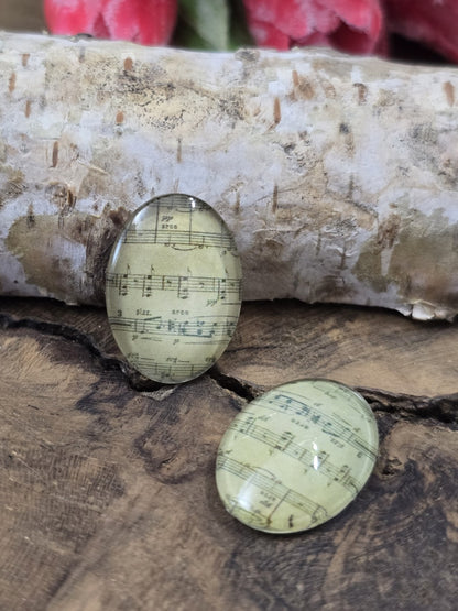 Zwei ovale Glascabochons (18x13 mm) mit antikum Notenblatt-Motiv dekorativ platziert auf rustikalem Holz vor einem hellen Birkenstamm.