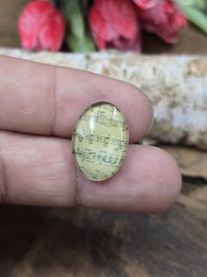 Ein ovaler Noten-Glascabochon (18x13 mm) im Vintage-Musik-Design auf einer Fingerspitze als praktischer Größenvergleich für DIY-Schmuck.